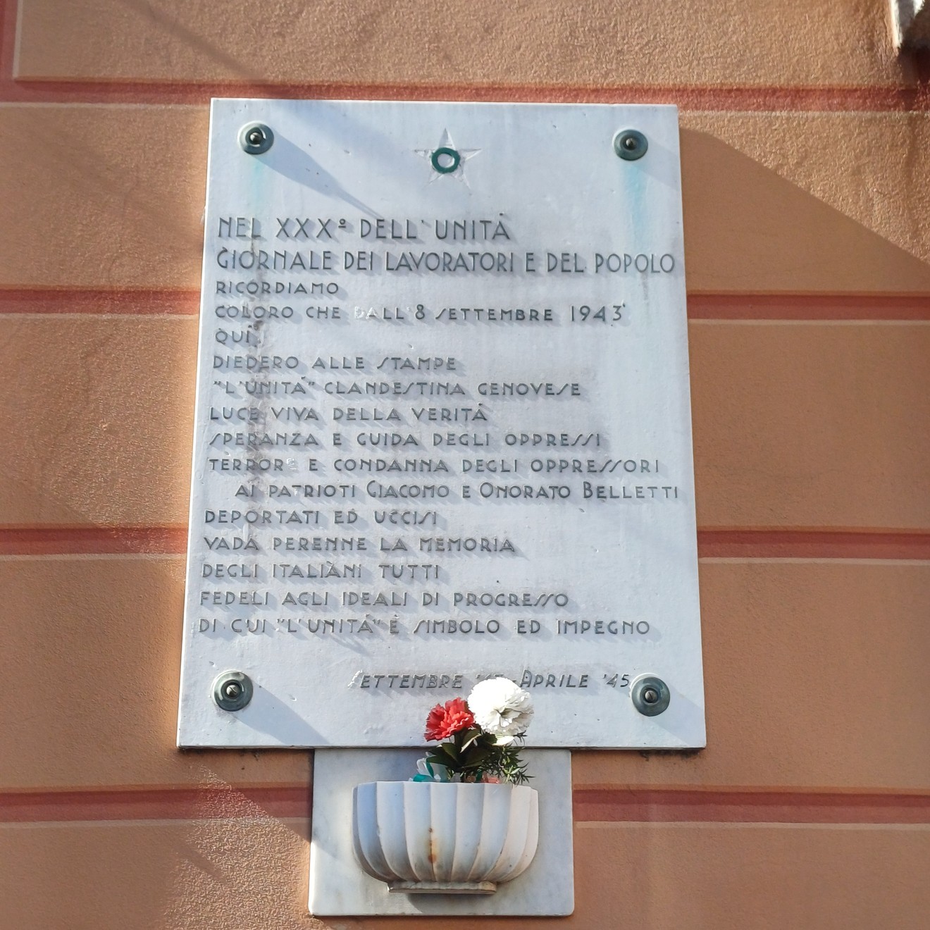 Lapide in marmo con iscrizione su parete, sotto un piccolo vaso con fiori.