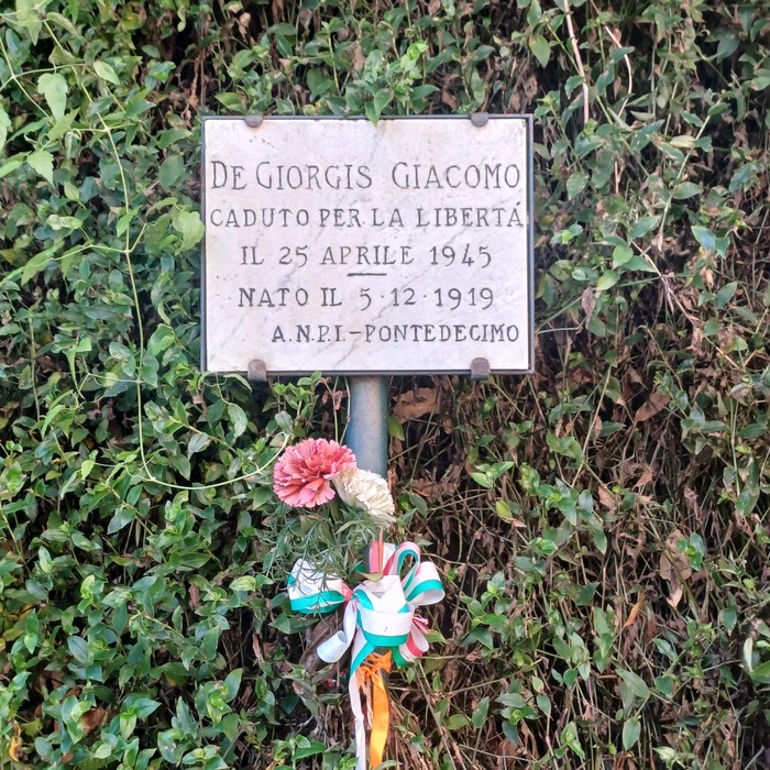 Targa in marmo con iscrizione, fissata su un palo tra la vegetazione, con garofani e coccarda tricolore.