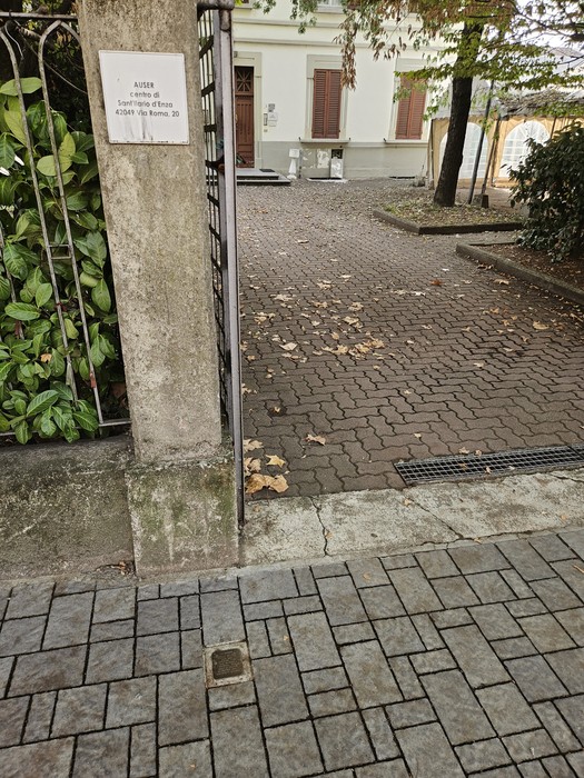 Pilastro in pietra con targa e cancello in ferro, all'ingresso di un vialetto pavimentato a ciottoli.