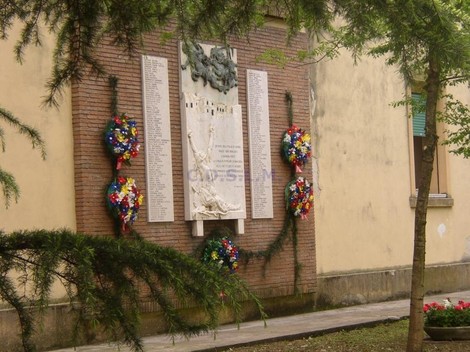 Lapide per le vittime dei bombardamenti, adornata con fiori commemorativi, con un muro di mattoni sullo sfondo.