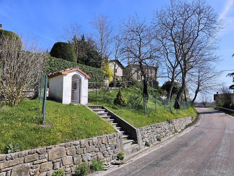 Piccola cappella bianca con portale ad arco, su un pendio erboso lungo una strada di collina.