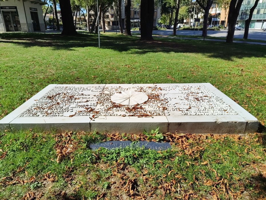 Lastra orizzontale in pietra con iscrizione incisa e decoro centrale, posata su prato tra alberi.