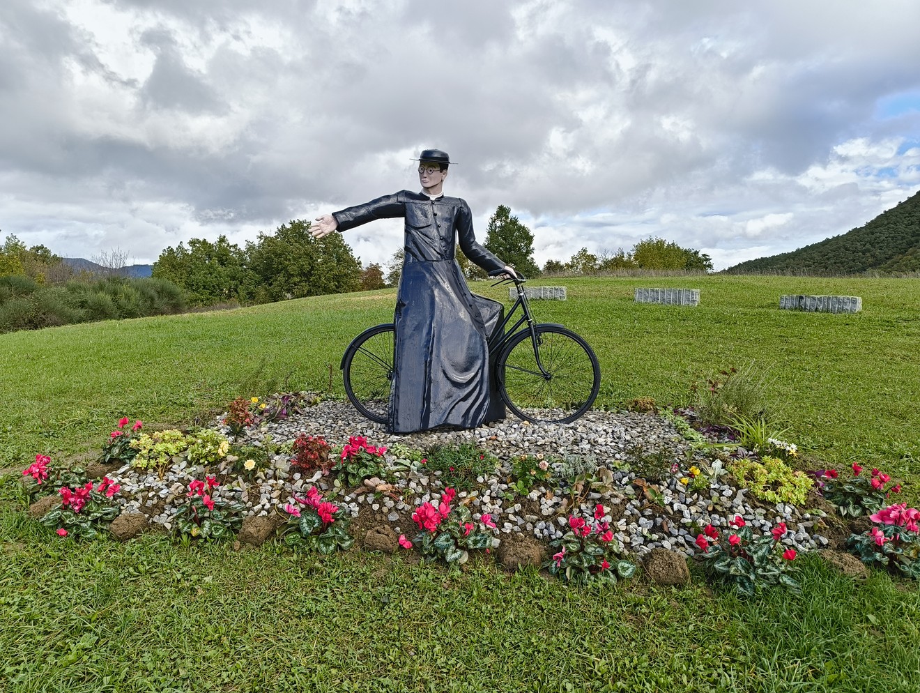 Statua di un angelo su una bicicletta circondata da fiori, in un contesto naturale con cielo nuvoloso.