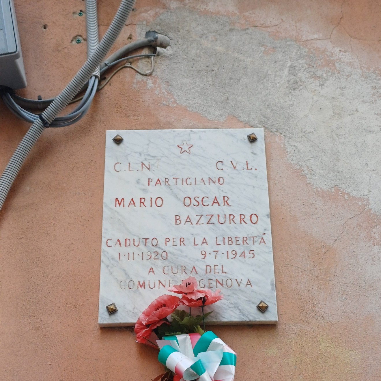 Lapide in marmo su parete esterna, con iscrizione e fiori deposti alla base.