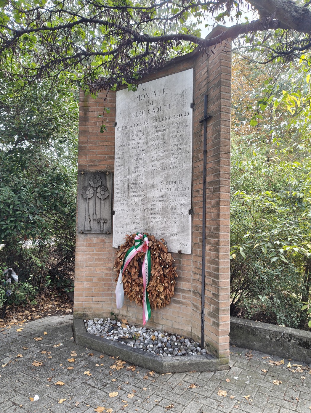 Monumento in mattoni con lapide incisa, decorazione in ferro battuto e corona con nastro tricolore, tra gli alberi.