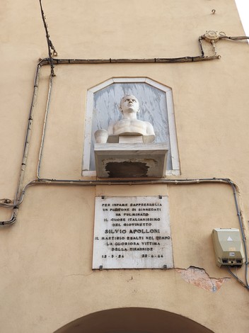 Lapide commemorativa con busto di Apolloni, affissa a una parete, in un contesto urbano.
