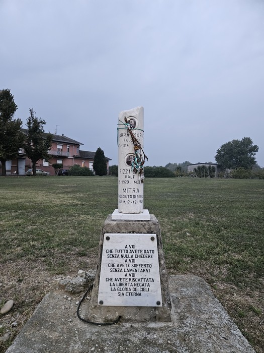 Cippo in pietra su basamento, con fotografie e iscrizioni, in un prato erboso.