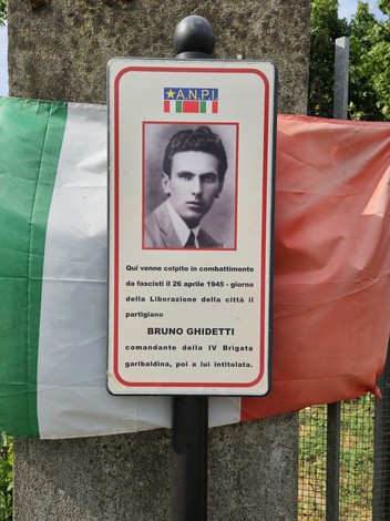 Croce commemorativa dedicata a Bruno Ghidetti, partigiano caduto nel 1945, con bandiere italiane sullo sfondo.