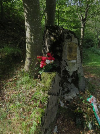 Monumento della fossa comune con fiori rossi, immerso nella vegetazione e circondato da alberi.