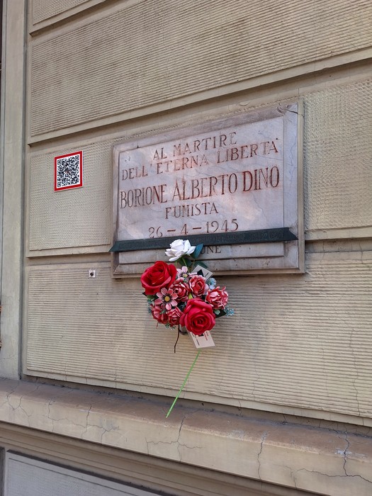 Lapide in marmo su parete esterna, con iscrizione, fiori rossi e bianchi deposti sotto.