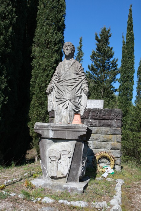 Monumento ai caduti di Purocielo, statua in pietra con dettagli e alti cipressi sullo sfondo.