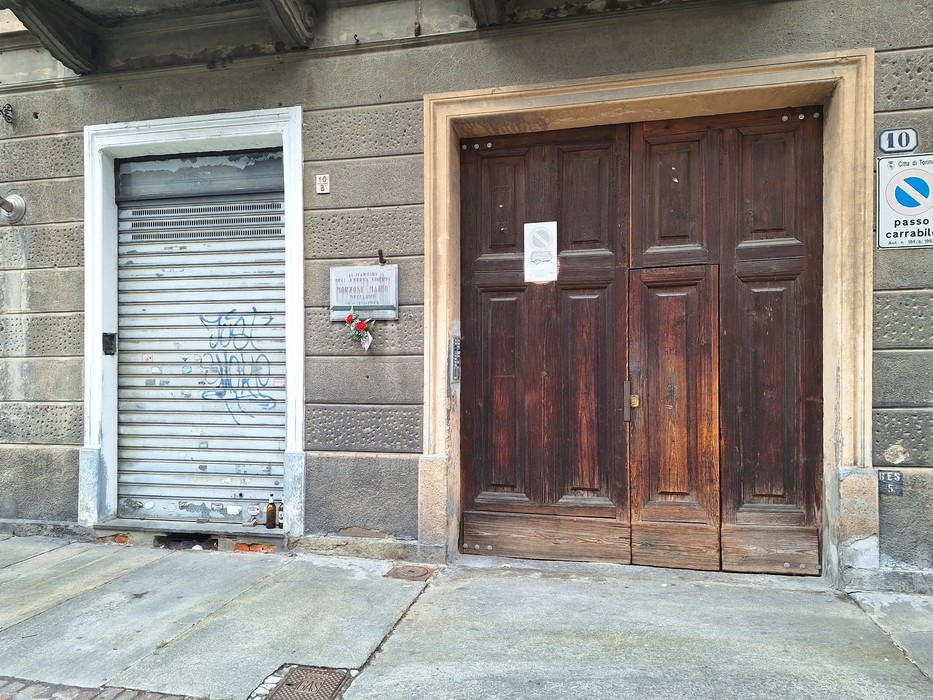 Lapide commemorativa su un muro accanto a un'entrata con porte di legno e un serramento grigliato.