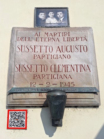 Lapide in marmo su parete, con iscrizione e foto di tre persone incorniciata sopra, lampada votiva sotto.