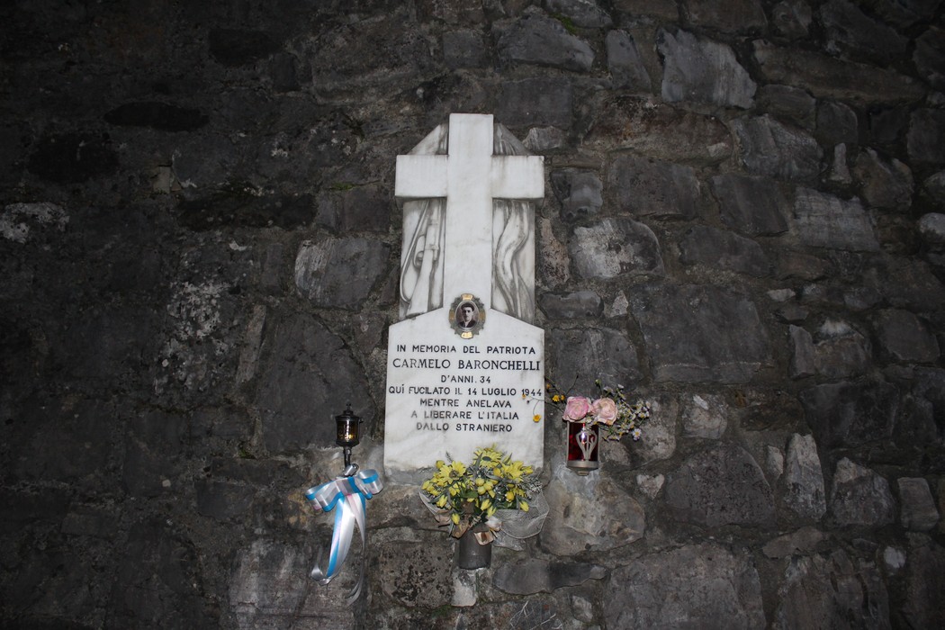 Lapide commemorativa di Carmelo Baronchelli, con fiori e una croce, situata su una parete di pietra.