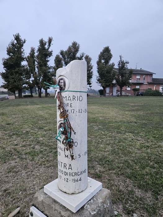 Cippo in marmo bianco con iscrizione e foto, su prato alberato, con fiori e oggetti votivi appesi.