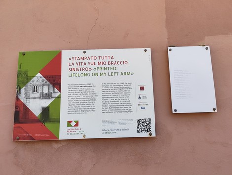 Due targhe su parete rosa, una colorata con iscrizione bilingue, l'altra bianca con testo e QR code.