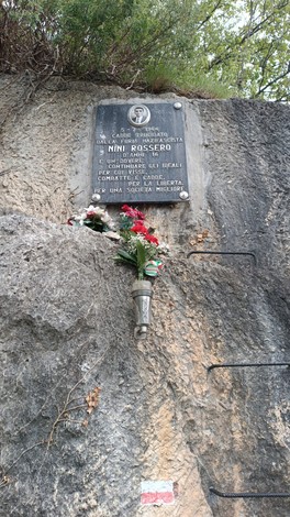 Lapide commemorativa dedicata a Nini Rossero, collocata su una roccia con fiori e un candeliere.