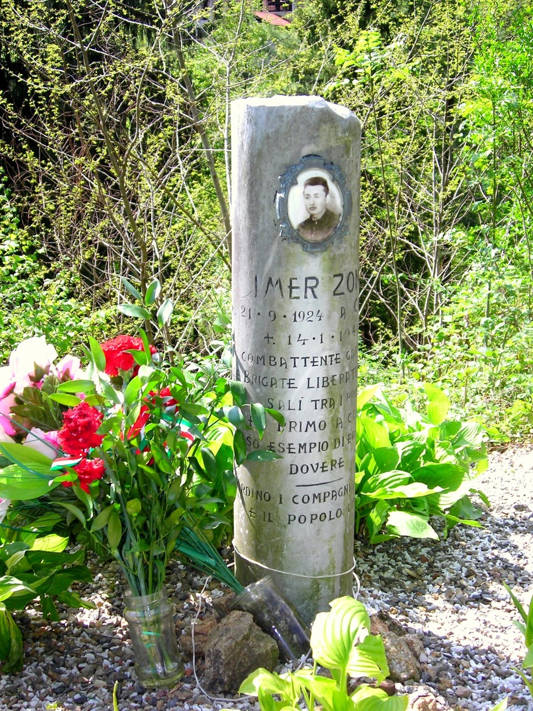 Cippo commemorativo di un combattente, circondato da fiori, in un contesto naturale.