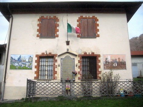Lapide commemorativa ai caduti di Valle, adornata da fiori e bandiere, su un edificio storicamente significativo.