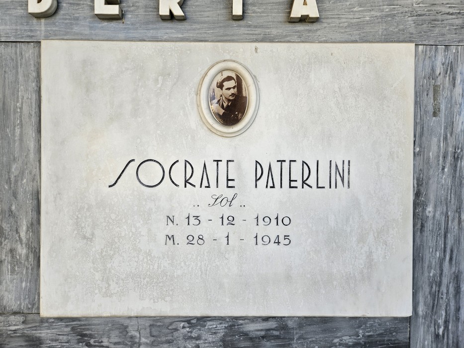 Lapide in marmo con ritratto ovale fotografico e iscrizione dedicata a un caduto, fissata su parete esterna.
