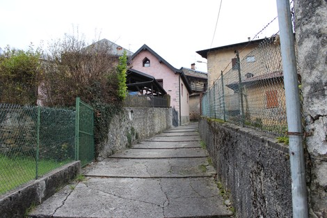 Strada in salita con case storiche ai lati, circondata da vegetazione e recinzioni. Atmosfera tranquilla e rurale.