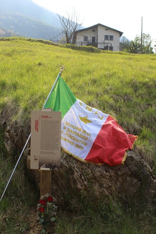 Impronta partigiana con bandiera italiana e targa commemorativa in un paesaggio montano.