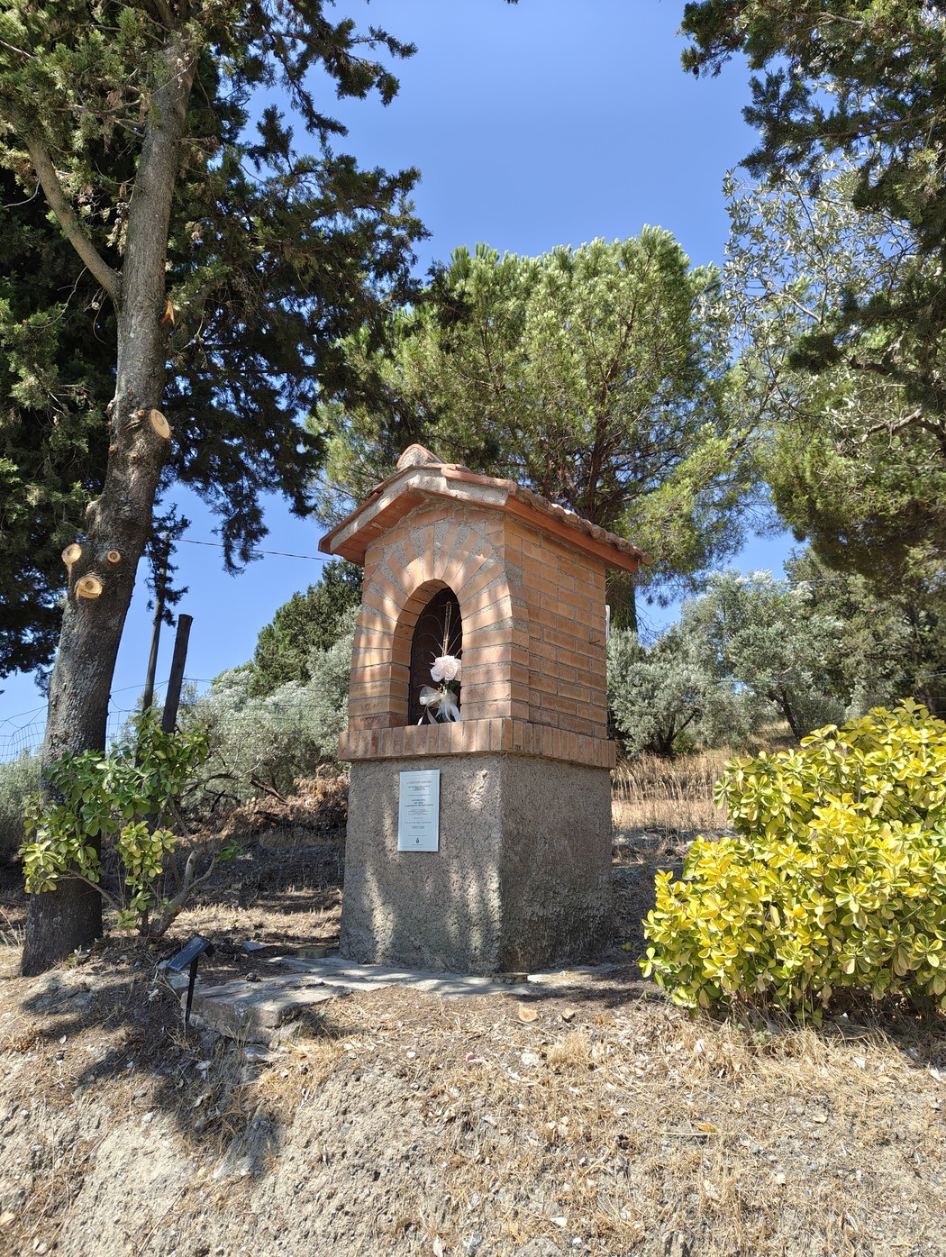 Tabernacolo in mattoni con nicchia ad arco e tetto spiovente, base in pietra con lapide, tra alberi.