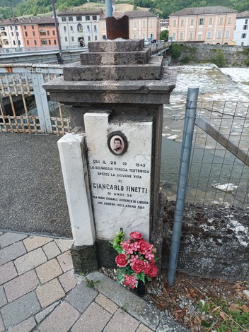 Cippo commemorativo di Cianalbi Finetti, con fiori e foto, sullo sfondo il fiume e case storiche.