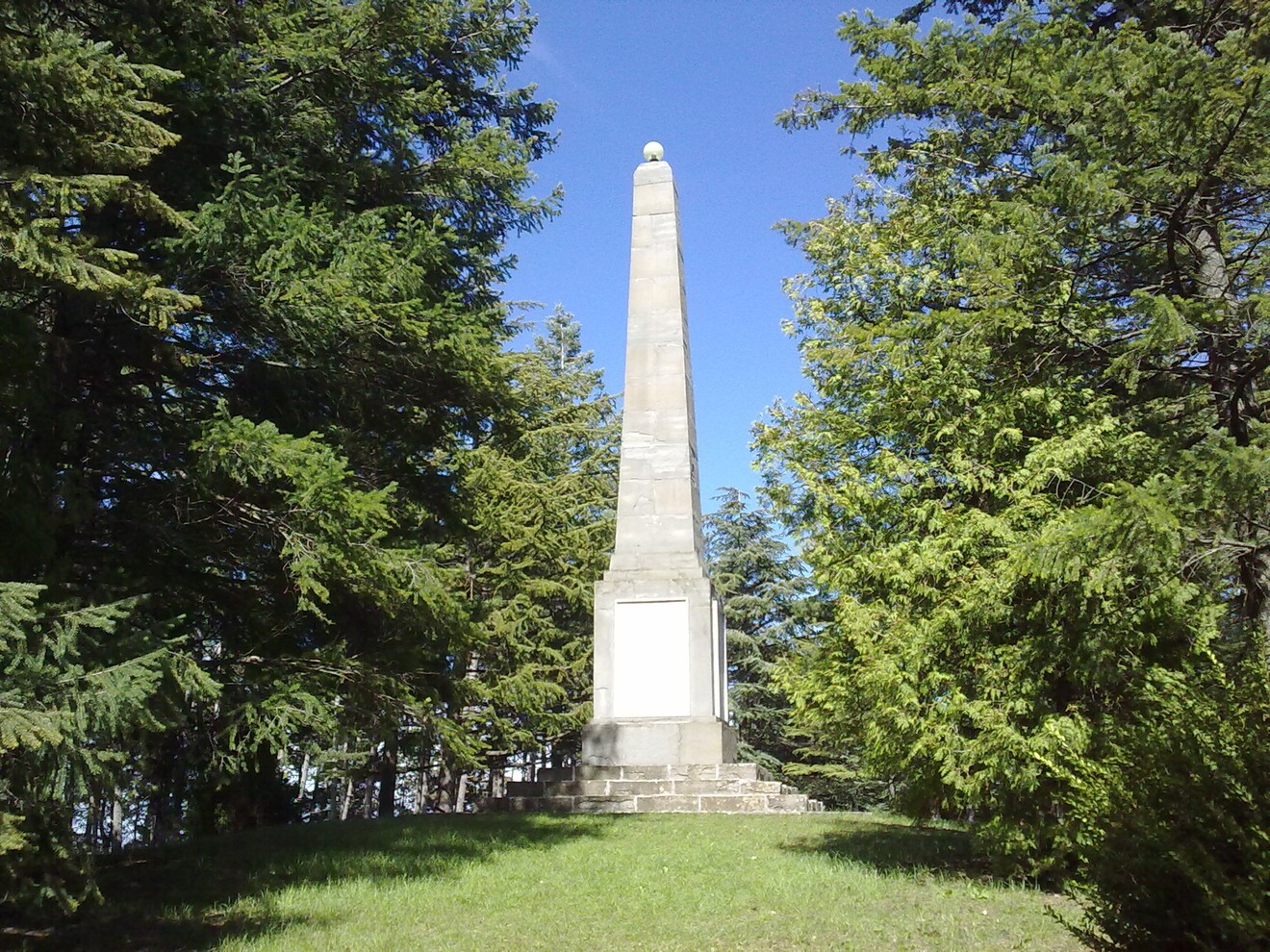 Monumento ai Caduti di Castelnovo, obelisco circondato da alberi in un paesaggio sereno.