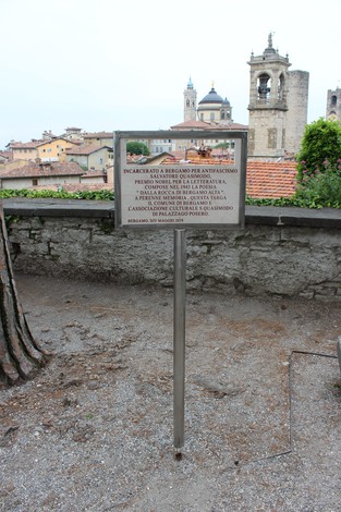 Targa commemorativa dedicata a Quasimodo, con vista su un paesaggio urbano e edifici storici.