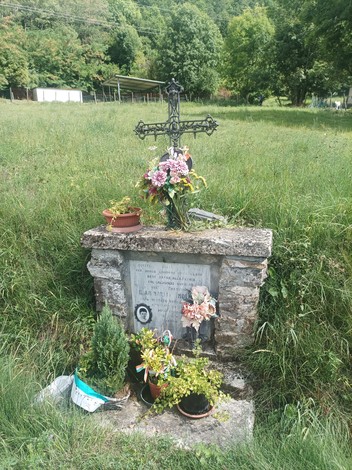 Cippo commemorativo con croce, fiori e piante, immerso nella natura verde.