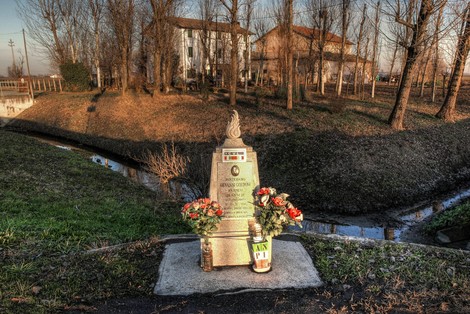 Cippo commemorativo dedicato a Goldoni, circondato da fiori e vista del paesaggio circostante.