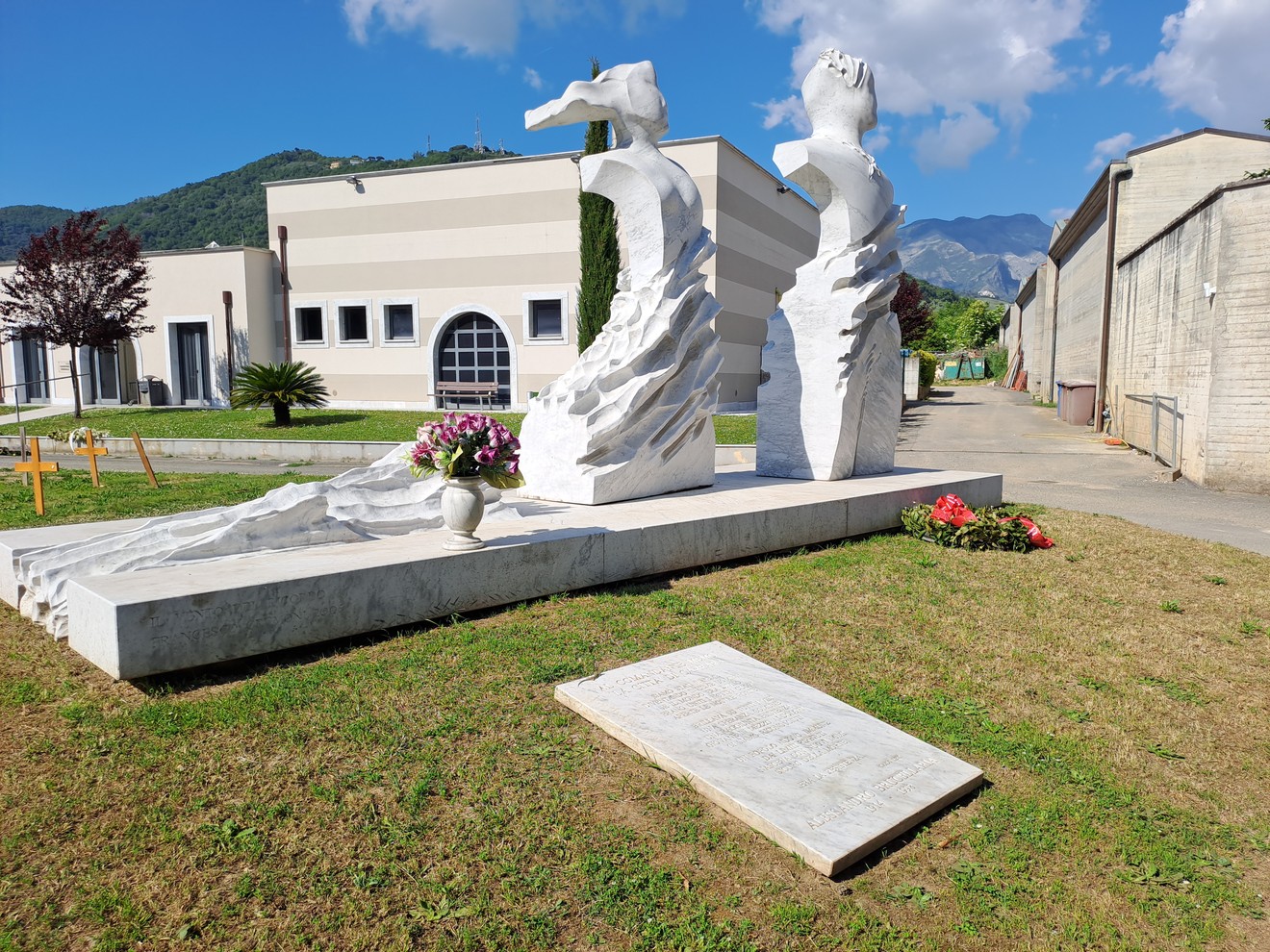 Monumento a Brucellaria con sculture di marmo e dettagli floreali in un'area verde. Sfondo montuoso e cielo sereno.