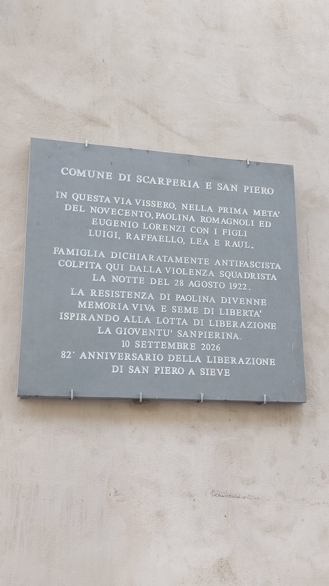 Lapide in pietra grigia con iscrizione, affissa su una parete esterna intonacata.