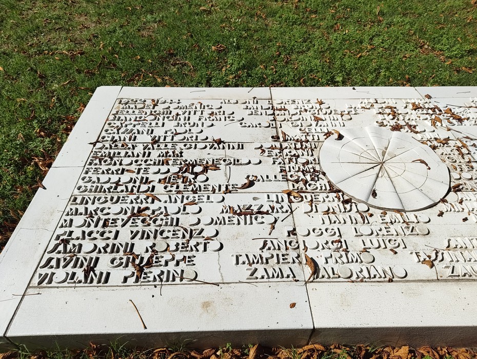 Lapide in pietra bianca a terra, con lunghi elenchi di nomi incisi, posata sul prato.