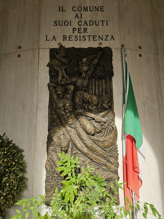 Scultura commemorativa al "Sacrario dei Caduti per la Resistenza" con bandiera italiana e vegetazione circostante.