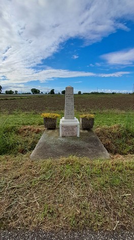 Cippo commemorativo incastonato in un paesaggio rurale, circondato da fiori e cielo blu.