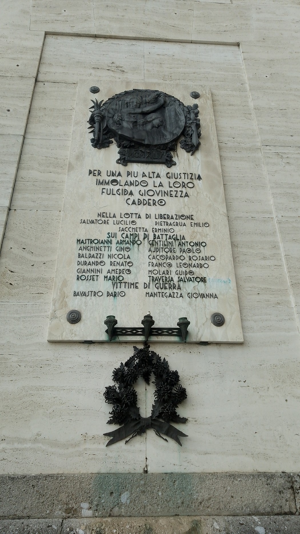 Lapide in marmo con iscrizione e bassorilievo, affissa a una parete, con corona in metallo sottostante.