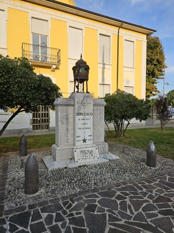 Monumento in pietra con iscrizione e fregio bronzeo, su piazza con proiettili ai lati, davanti a un edificio giallo.