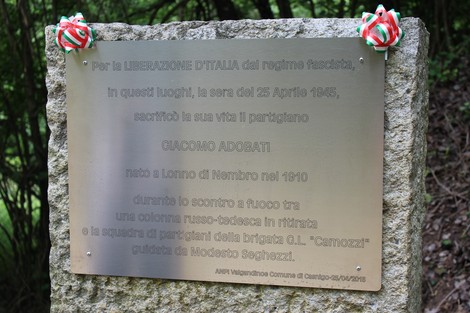 Cippo commemorativo dedicato a Giacomo Adobati, situato in un'area verde, con decorazioni tricolori.