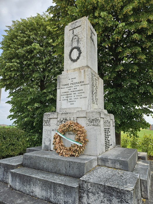 Monumento ai Caduti con corona commemorativa e monumento di pietra, circondato da alberi e verde.