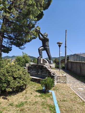 Statua in bronzo di un uomo con braccia alzate, su basamento in pietra, tra alberi e vegetazione.