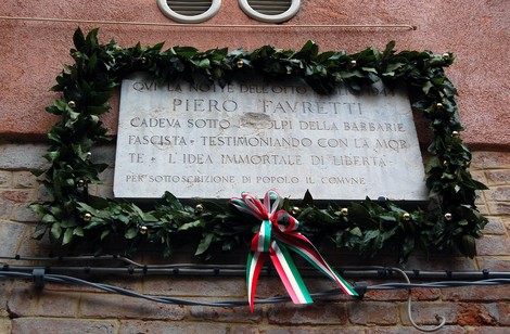 Lapide commemorativa dedicata a Piero Favretti, circondata da una coroncina di foglie e decorazioni colorate.