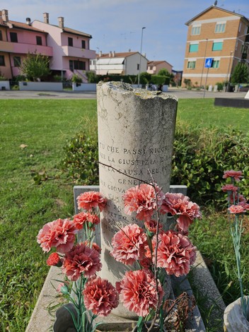 Cippo commemorativo con fiori rossi in un'area verde, circondato da edifici residenziali.