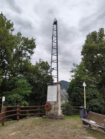 Un faro monumentale eretto su una collinetta, circondato da alberi e con un paesaggio montano sullo sfondo.