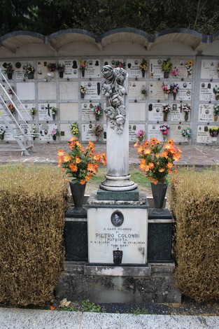 Cippo funerario dedicato a Pietro Colombi, con statuetta e fiori artificiali, in un cimitero.