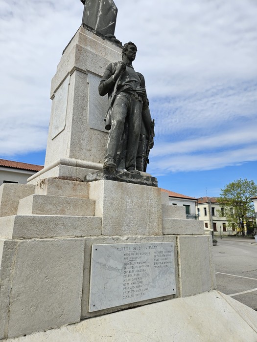 Monumento ai Caduti di Grignano Polesine, con statua e base in pietra, circondato da edifici e cielo sereno.