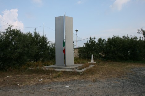 Monumento commemorativo con obelisco e bandiera italiana, immerso in un'area verde.