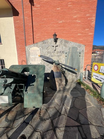 Monumento in pietra con croce e iscrizione, elica di aereo e cannone davanti a una parete in mattoni.