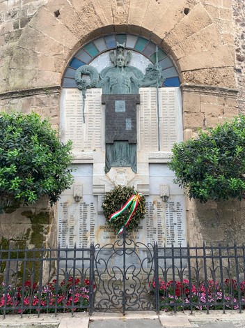 Monumento ai Caduti di Alghero, con statue e elenchi commemorativi, circondato da piante e fiori.